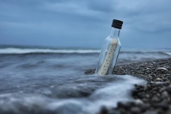 A "message in a bottle" sat on a shingle beach with the waves lapping the base of the bottle.